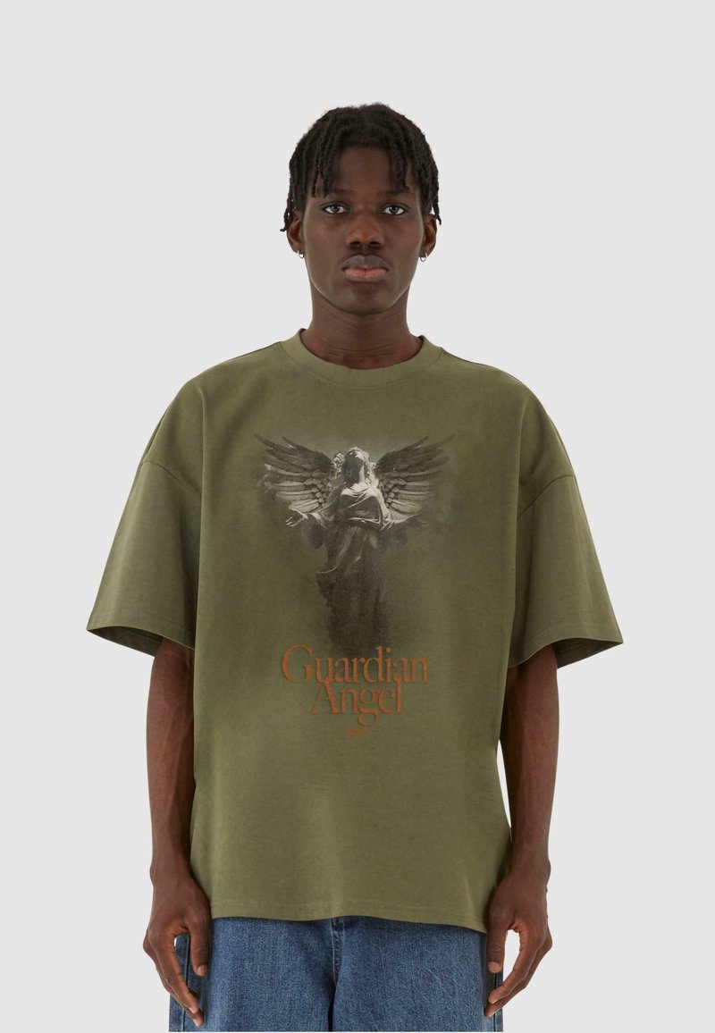 Olivenfarvet oversized T-shirt med et centralt grafisk tryk af en skytsengel og tekst nedenfor, lavet af tekstureret bomuld med korte ærmer.