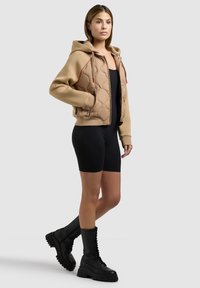 Femme portant un sweat à capuche matelassé beige, des cuissards noirs et des bottes de combat noires à lacets, debout les mains dans les poches.
