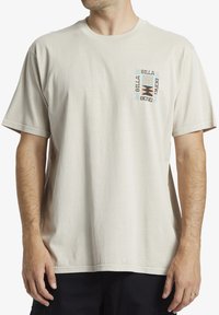 Camiseta de manga corta color beige hecha de algodón, con un logo gráfico en negro y azul en el lado izquierdo del pecho. Ajuste casual y relajado.
