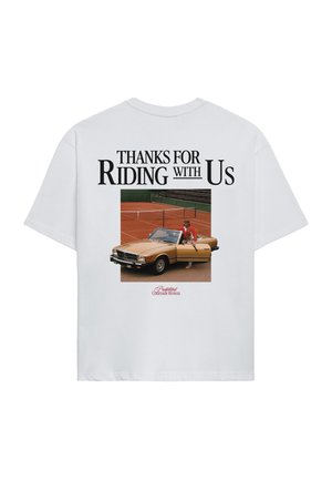 Young man wearing white oversized T-shirt with red text, black patterned neck scarf, green baseball cap with orange logo, and gray pants.