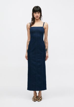 Robe maxi en denim bleu foncé avec un décolleté carré, des bretelles fines, des coutures verticales et une silhouette ajustée ; portée avec des chaussures à imprimé léopard.