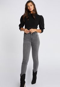 Top nero con maniche a sbuffo abbinato a jeans grigi a vita alta e slim-fit; modello che indossa stivali neri alla caviglia; sfondo semplice.
