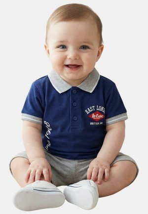 Bébé souriant assis jambes croisées, portant un polo bleu marine avec col et poignets gris, un short gris et des chaussures blanches.