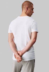 T-shirt en coton blanc à manches courtes, col rond et texture lisse. Le modèle se tient avec les mains jointes derrière le dos, portant un pantalon gris.
