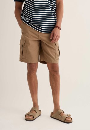 Mees, keda kannab pruunid cargo-lühikesed püksid, beežid sandaalid ja tumesinise-valge triibuline särk, seisab neutraalse tausta ees.