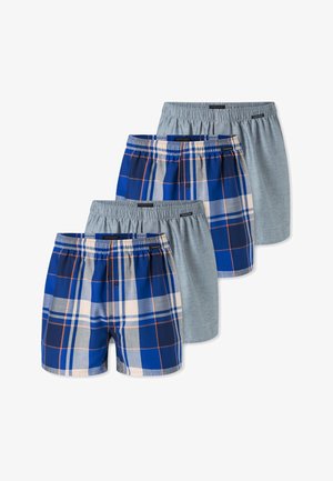 Muskelöser junger Mann mit kurzen Haaren, der blaue und beige karierte Boxershorts trägt und mit erhobenen Armen vor einem schlichten Hintergrund posiert.