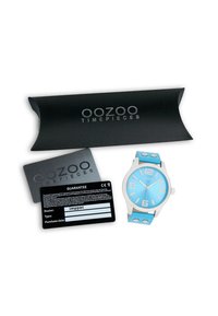Montre bleu clair avec un boîtier en argent et de grands chiffres blancs, assortie à un bracelet en silicone texturé. Comprend une carte de garantie noire et un emballage.