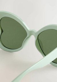 Occhiali da sole verde menta con montatura a forma di cuore, lenti verde scuro e aste sottili e curve. Materiale plastico liscio.
