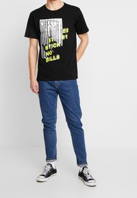 Sorte bomulds t-shirt med grafisk tekst i hvid og gul. Korte ærmer og rund hals. Bæres med blå jeans og sorte sneakers.