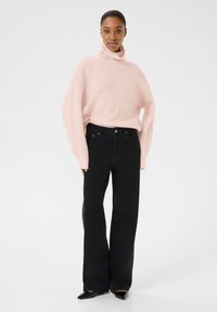 Prevelik roza pulover s turtleneck ovratnikom in dolgimi rokavi, v kombinaciji z črnimi širokimi kavbojkami. Gladka tekstura, minimalistična zasnova in brez vidnih vzorcev.