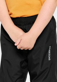 Mustat vedenpitävät housut, joissa on joustava vyötärö, ja sivussa valkoisella tekstillä merkin nimi "Didriksons". Sujuva pinta.