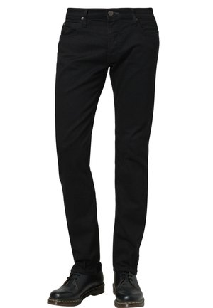 Jean en denim noir avec une coupe slim, comportant cinq poches, une fermeture par bouton sur le devant et des détails de couture visibles. Porté avec des chaussures noires.