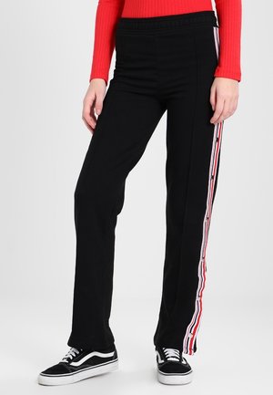 Sorte bukser med lige ben med røde, hvide og pink stribede trykknapper ned langs siderne, båret med sorte sneakers og en rød ribstrikket sweater.