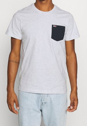 Homme portant un t-shirt gris clair à manches courtes avec une poche noire sur la poitrine et un jean bleu clair, debout les bras détendus le long du corps.