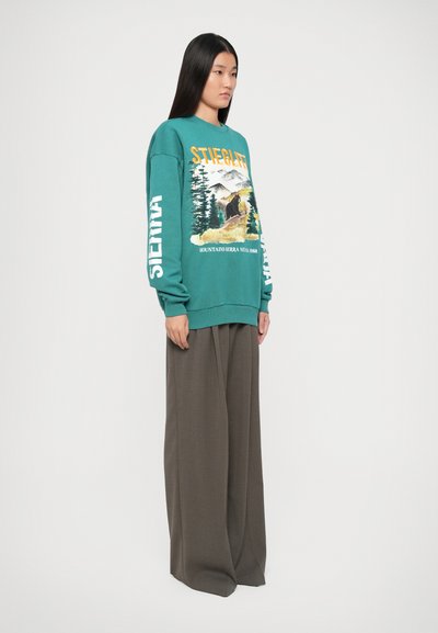 Felpa verde acqua con grafica di un orso e alberi; testo bianco sulle maniche. Abbinata a pantaloni larghi a vita alta di colore marrone scuro.