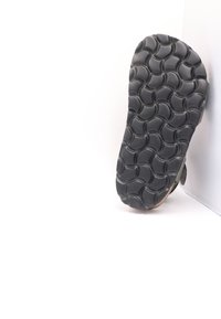 Semelle de chaussure en caoutchouc noir avec un motif ondulé et une surface texturée. Le bord montre des signes d'usure ; s'ajuste parfaitement contre un fond blanc.