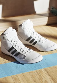 White athletic shoes made of mesh and synthetic material, featuring three black stripes, high top design, and a textured sole.