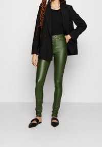 Kvinna i svart kavaj och topp, gröna högmidjade läderbyxor, svarta spetsiga ballerinaskor, med halsband och ring som accessoarer.