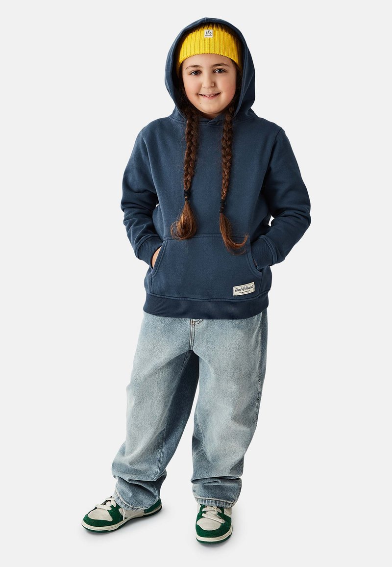 Laivastonsininen huppari kengurutaskulla ja resorireunuksilla, yhdistetty vaalean sinisiin löysiin farkkuihin ja vihreänvalkoisiin tennareihin. Keltainen neulottu pipo.