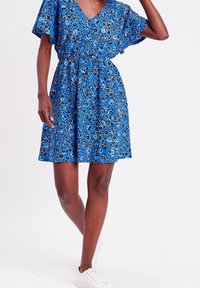 Blauwe bloemenjurk met korte mouwen, V-hals, een smalle taille en een uitlopend rokje, gecombineerd met witte sneakers. Het patroon bevat kleine zwarte bloemen.