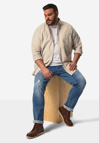 Camisa de rayas beige de manga larga sobre una camiseta blanca, combinada con unos vaqueros azules desgastados y botas marrones, sentado en un bloque de madera.
