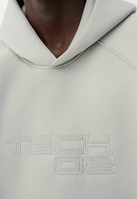 Ljusgrå huvtröja med en strukturerad yta, med en logotyp "TECH 02" i präglad vit broderi på framsidan, och en inhemsk huva design.
