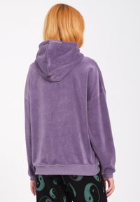 Sudadera morada de tela suave y aterciopelada con un corte relajado, que presenta una capucha grande y puños acanalados, combinada con pantalones negros con estampado.