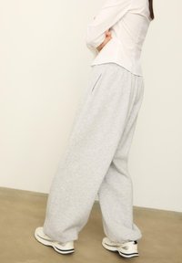 Graue Sweatpants mit lockerer Passform, die über Seitentaschen und einen elastischen Bund verfügen. Getragen mit weißen Turnschuhen vor einem neutralen Hintergrund.