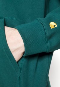 Felpa teal con polsini a coste e tasca frontale. Accentatura del logo dorato sulla manica. Texture liscia con dettagli di cucitura visibili.