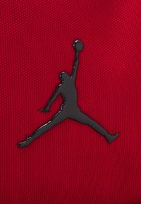 Piros textil háttér, amelyen egy kiemelkedő fekete Jumpman logó látható, amely egy ugrás közben lévő figurát ábrázol, sima textúrával és fényes felülettel.