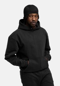 Schwarze Kapuzenpullover aus strukturiertem Stoff, mit einer Vordertasche und lockerer Passform. Getragen mit passenden schwarzen Hosen und einer eng anliegenden Mütze.