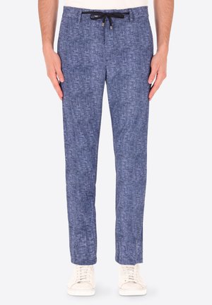 Uomo con pantaloni blu testurizzati con coulisse e scarpe da ginnastica bianche, in piedi dritto contro uno sfondo semplice.