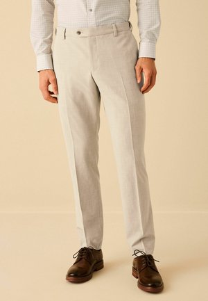 Pantaloni grigi chiari con una texture liscia, dotati di vita piatta, passanti per cintura e una vestibilità su misura. Indossati con scarpe in pelle marrone scuro.