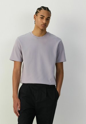 Ung mand med fletninger iført en lysegrå kortærmet skjorte og sorte bukser, stående mod en ensartet lys baggrund.