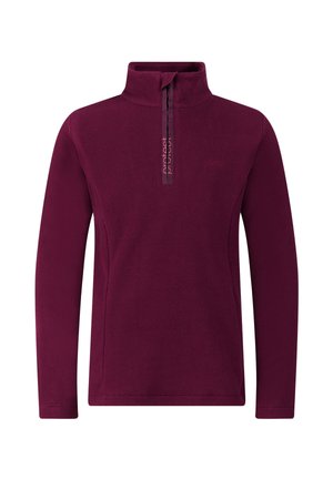 Jonge persoon die een blauwe muts verstelt, draagt een paarse lange mouwen met rits, beige broek en zwarte schoenen, staat tegen een effen achtergrond.