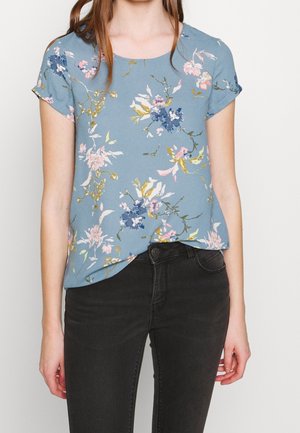 Femme portant un chemisier bleu à manches courtes fleuri, associé à un jean noir ajusté, debout devant un fond clair uni.