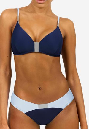 Frau trägt einen zweifarbigen Bikini mit navyblauen Cups und Höschen, hellblauen Trägern und Bund sowie grauen Akzenten vorne in der Mitte.