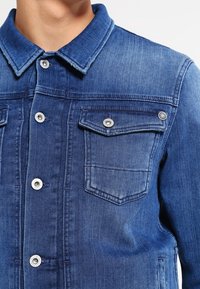 Chaqueta de mezclilla en azul oscuro con cuello, botones de presionar plateados, bolsillos en el pecho y una tela texturizada con un ligero efecto de desgaste.