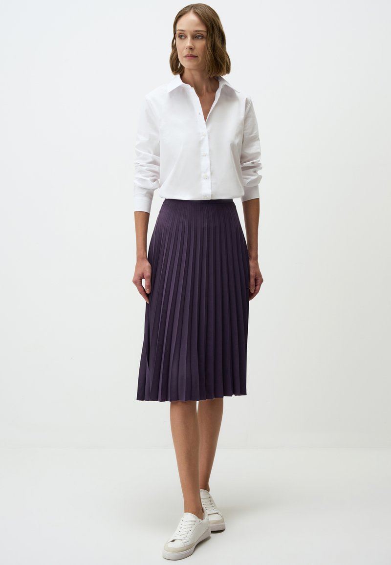 Camicia bianca con maniche lunghe, abbinata a una gonna plissettata viola scuro lunga fino al ginocchio e a sneaker bianche su uno sfondo neutro.