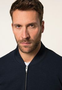 Chaqueta bomber con cremallera y cuello acanalado en color navy. El fondo es neutro, centrando la atención en la textura y el ajuste de la prenda.