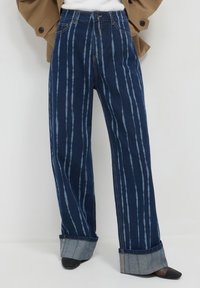 Jeans a gamba larga blu scuro con leggere righe verticali e ampi risvolti, indossati con scarpe nere a punta.