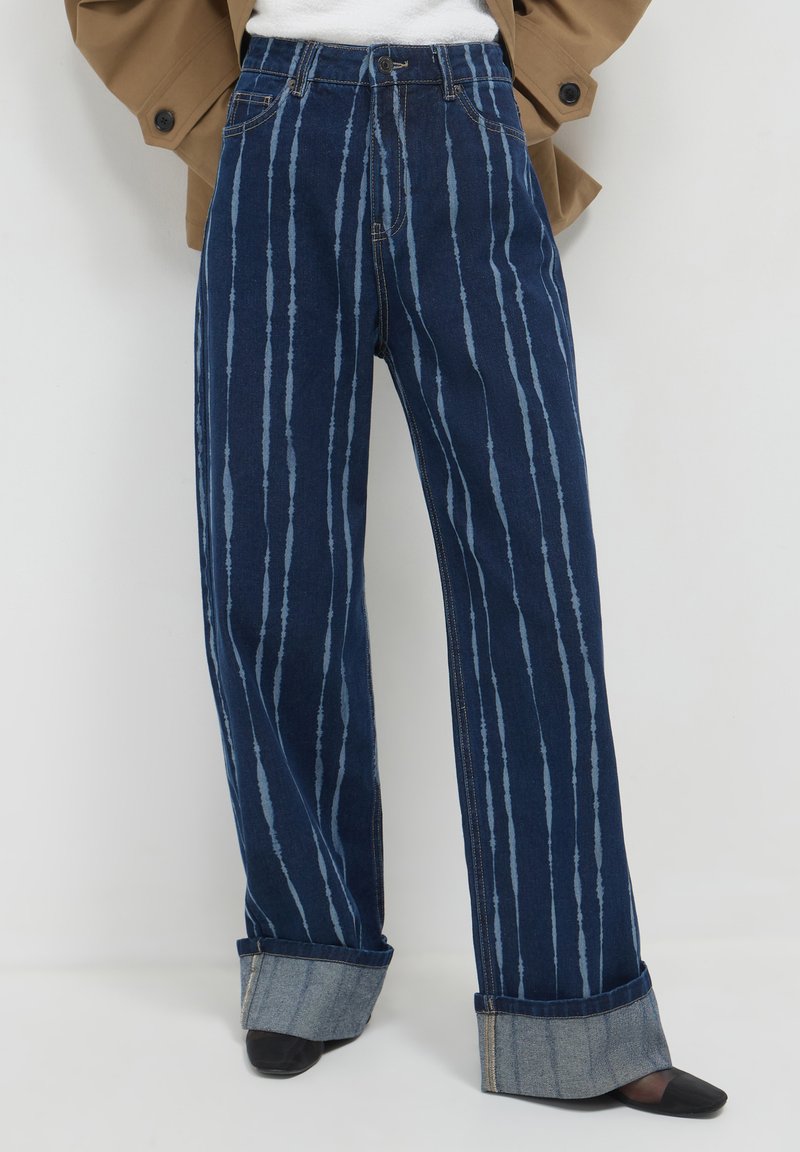 Jeans a gamba larga blu scuro con leggere righe verticali e ampi risvolti, indossati con scarpe nere a punta.