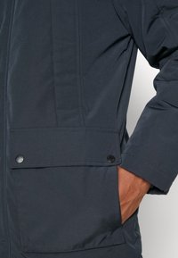 Donkerblauwe jas met een grote voorvakklep die wordt vastgehouden door twee zwarte knopen, met een hand gedeeltelijk in het vak aan de rechterkant.