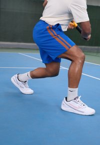 Sinised sportlikud lühikesed püksid pruunide triipudega, valged tennisetossud ja valged pahkluu sokid, paigutatud sinisele tennis courts. Keskenduge liikumisele.