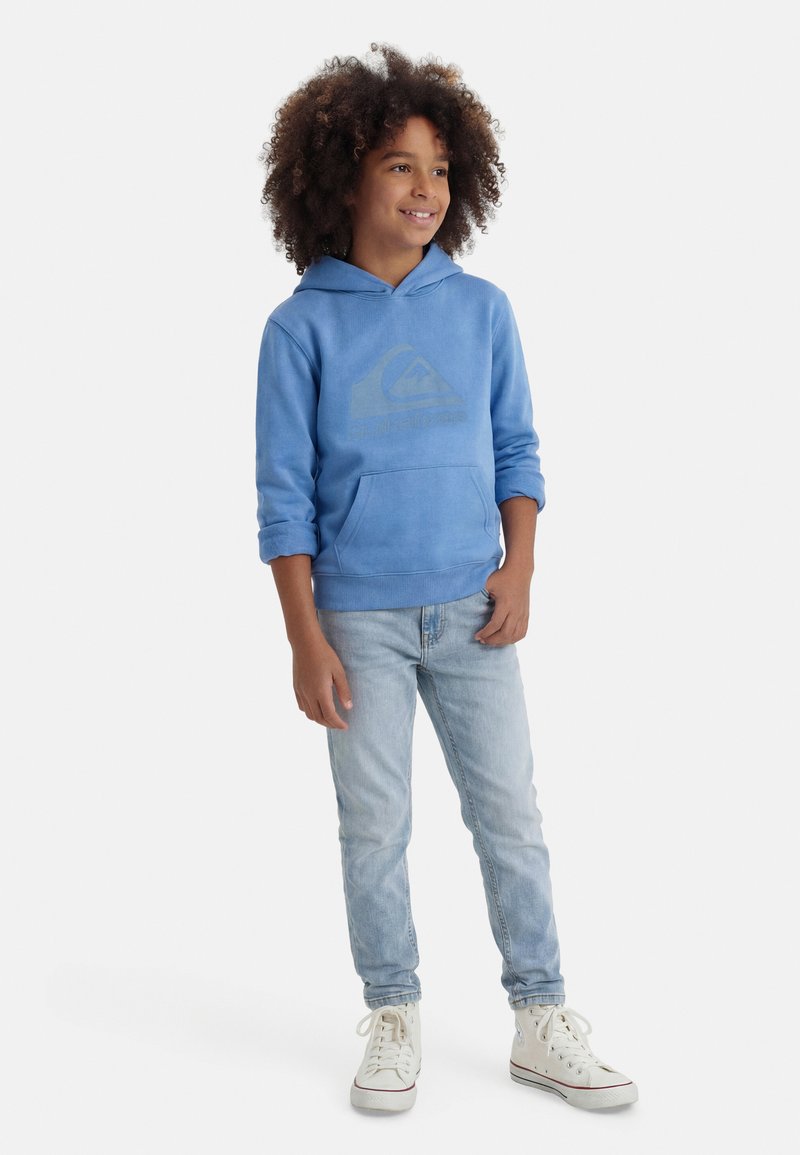Niño con cabello rizado vistiendo una sudadera con capucha azul, jeans claros y zapatillas blancas de caña alta, de pie con una postura relajada y sonriendo.