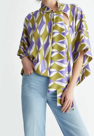 Kobieta ubrana w luźną bluzkę z abstrakcyjnym wzorem liści w kolorach oliwkowej zieleni, lawendowym i białym, w połączeniu z jasnoniebieskimi dżinsami, ręka w kieszeni.