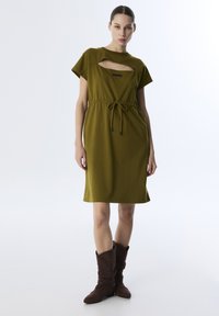 Olivgrünes Kleid mit runder Ausschnitt und Cut-Out-Details, an der Taille mit einem Kordelzug gerafft. Das Modell trägt braune Wildlederstiefel.