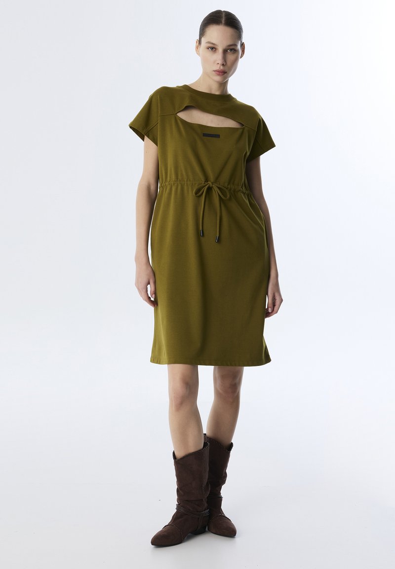 Olivgrünes Kleid mit runder Ausschnitt und Cut-Out-Details, an der Taille mit einem Kordelzug gerafft. Das Modell trägt braune Wildlederstiefel.