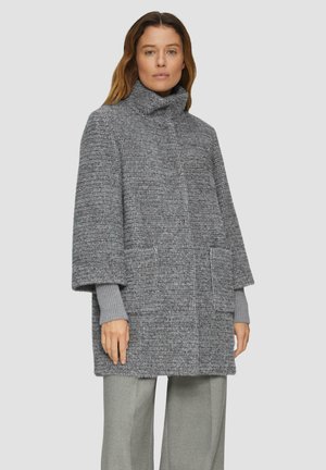 Manteau oversized en texture grise avec un col haut, deux poches avant et de larges manches 3/4, porté sur un haut gris clair et un pantalon assorti.
