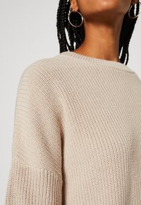 Gros plan d'une personne portant un pull en maille texturée beige, avec des cheveux tressés et de grandes boucles d'oreilles visibles.